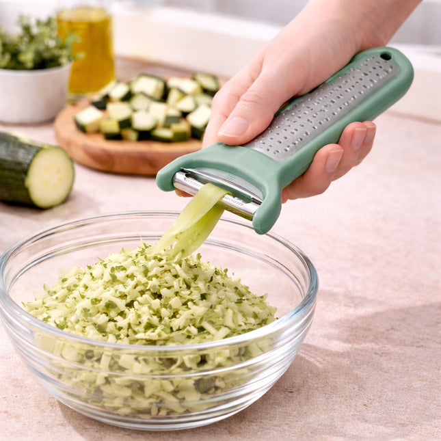 Person using a green vegetable grater to shred zucchini into a glass bowl on a kitchen counter.
