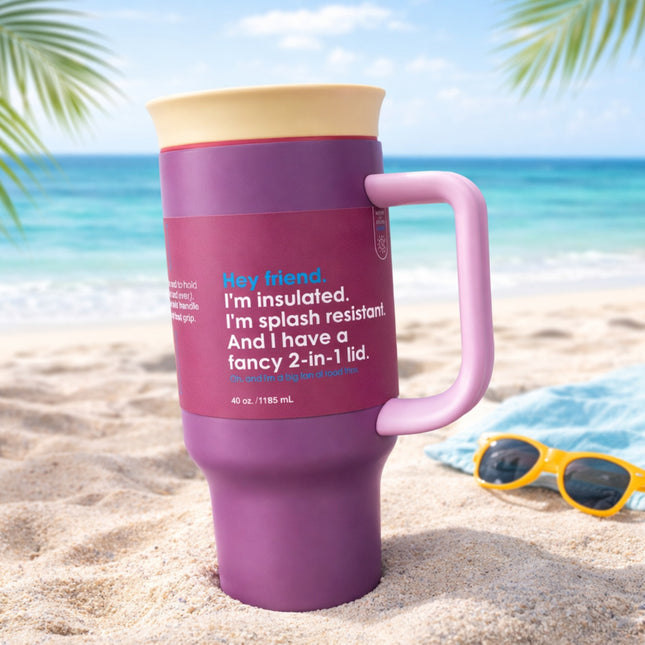 Purple travel mug with pink handle on a sandy beach with ocean and palm leaves in the background