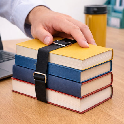 Stack of books tied together with a black strap on a wooden surface, with a hand placing another book on top.