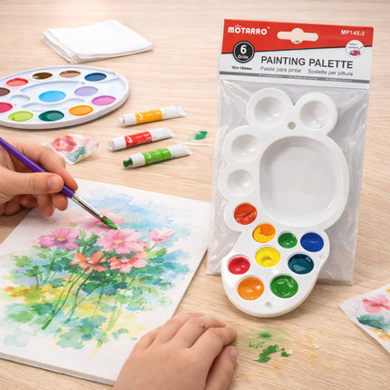 Person painting flowers with a watercolor palette and paintbrush on a wooden table.