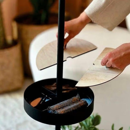 A close-up of a black metal candle holder with two light wood fan-shaped trays and two rolled candles, shown with hands adjusting the trays in a softly lit indoor setting.
