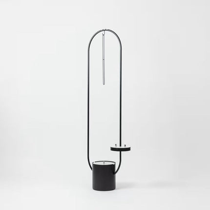 A modern black floor lamp with a cylindrical base, a thin curved metal frame arching over the top, and a small circular shelf, shown on a clean white background.