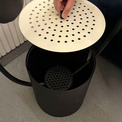 A close-up of a black cylindrical container with a round off-white perforated lid being lifted by a hand, revealing a black strainer basket inside.