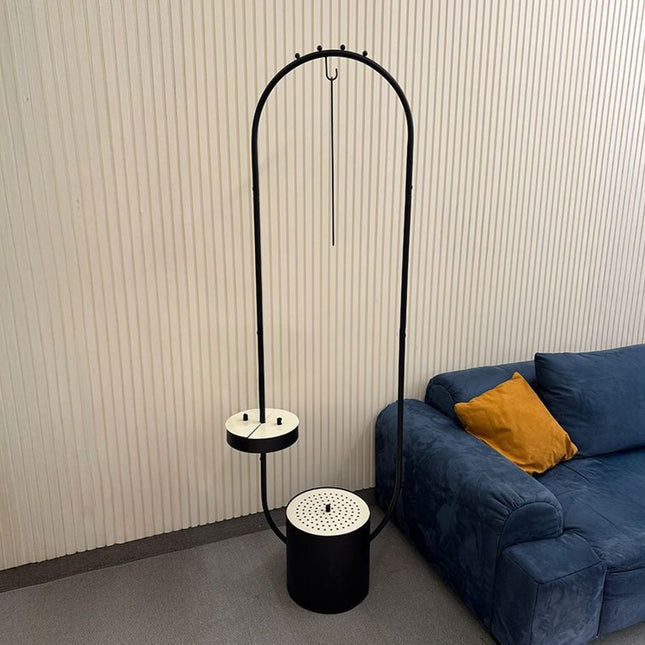 A black metal floor lamp with an arched top bar, a small circular shelf, and a cylindrical base, shown in a room next to a blue sofa with a yellow pillow.