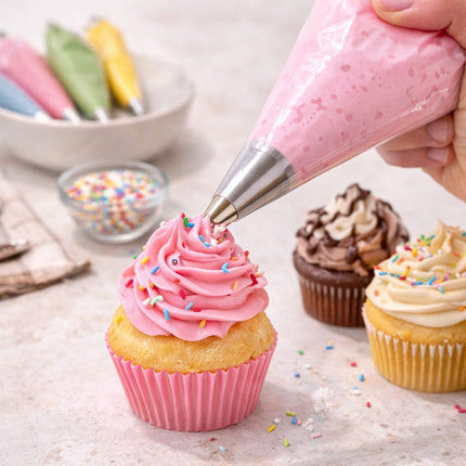 Person decorating a cupcake with pink frosting and sprinkles on a light surface.