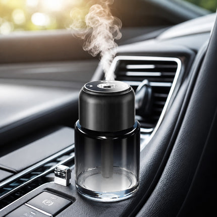 A black and clear car air freshener diffuser with a glass bottle and a small USB charging stick, shown on a car dashboard with white mist rising from the top.