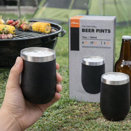Black insulated beer pints with silver rims held by a hand, with a box of beer pints and a grill in the background.