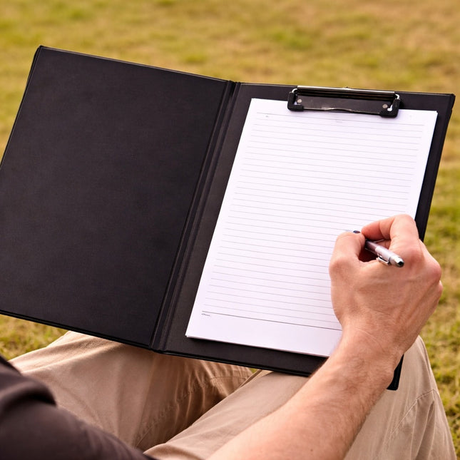 Person sitting outdoors with a black clipboard and notepad, writing with a pen.