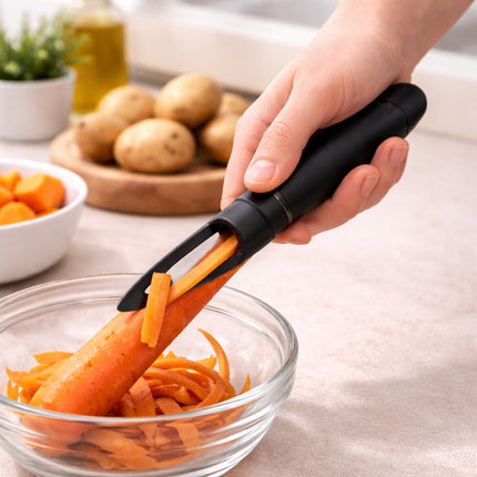 Person using a black vegetable peeler to peel a carrot over a glass bowl on a kitchen counter.