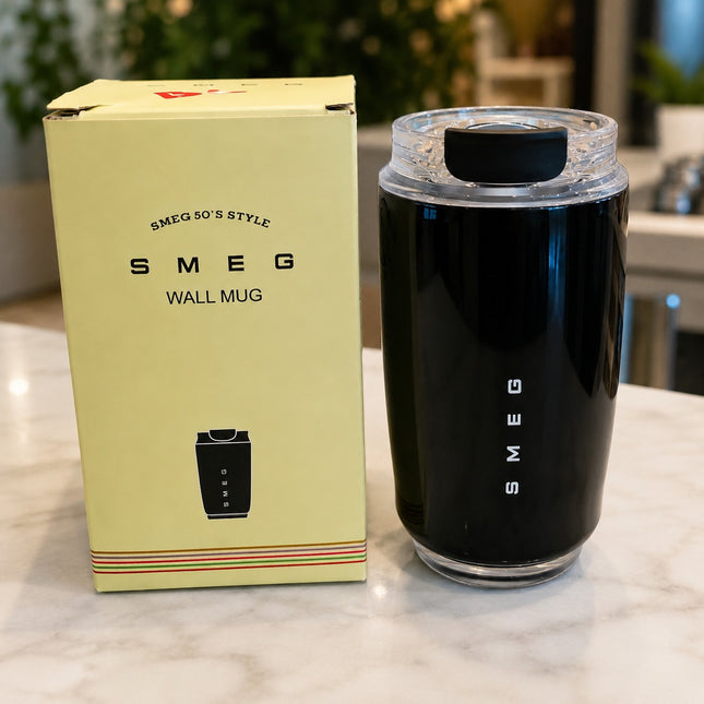 A black Smeg wall mug with a clear lid sits next to its pale yellow packaging box on a marble countertop.