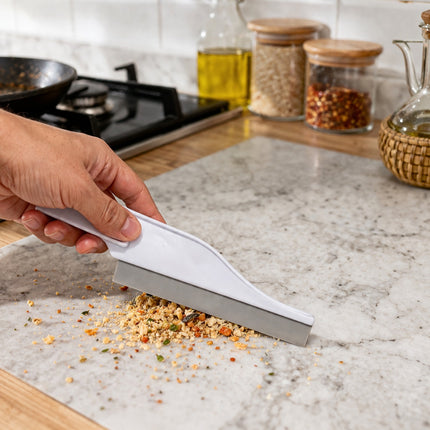 Person using a kitchen tool to collect crumbs on a marble countertop with a stove and jars in the background.