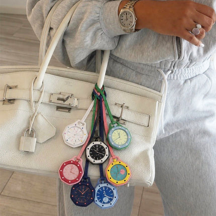 A person in a light gray sweatshirt carries a large off-white handbag with several colorful wristwatch charms hanging from the handle. The watches have round colorful faces and colorful straps, including pink, black, green, blue, yellow, and red.