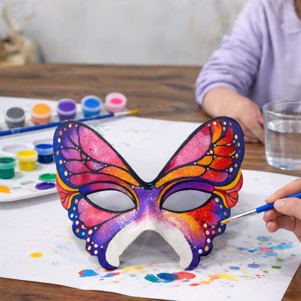 Colorful butterfly mask being painted on a table with art supplies.