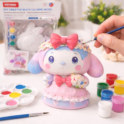 Colorful cartoon figurine with a hand painting it, surrounded by art supplies on a table.