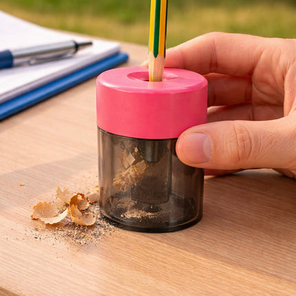 Hand using a pink pencil sharpener with a pencil inside on a wooden surface.