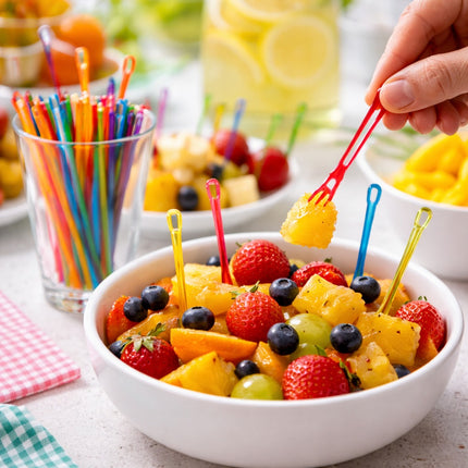 Fruit salad in a bowl with colorful skewers, surrounded by more fruit and drinks.