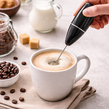Person using a milk frother to create foam in a cup of coffee on a table with coffee beans and milk.