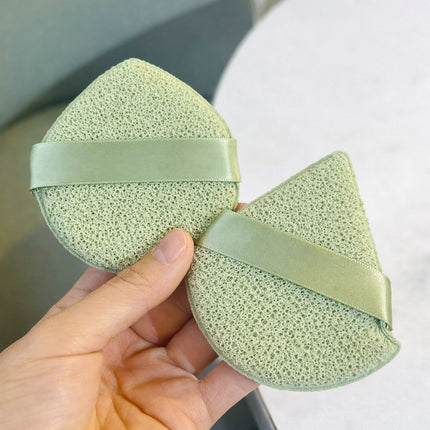 Two green makeup sponges with ribbons held by a hand against a neutral background