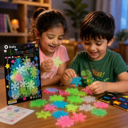 Two children, a girl in a pink shirt and a boy in a green dinosaur t-shirt, happily playing with colorful plastic snowflake toys on a wooden table. The product packaging by Italo is visible on the left, along with loose multicolor snowflakes and a sticker sheet.