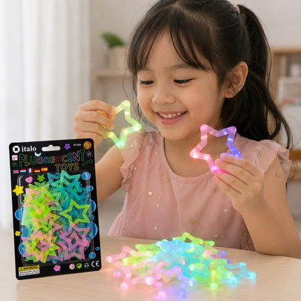 A young girl smiling while holding two glowing star-shaped toys, with more colorful fluorescent star toys scattered on a table in front of her. The package of Italo brand fluorescent toys is visible on the left.