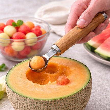 Hand using a melon baller to scoop out pieces of cantaloupe from a halved melon with a bowl of watermelon balls in the background.