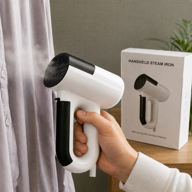 A hand holding a white and black handheld steam iron against a light-colored shirt, with the product box standing on a wooden table in the background.