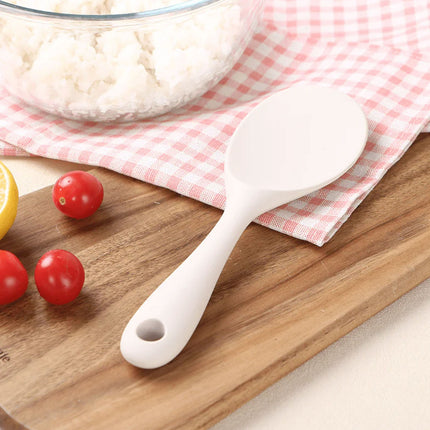 White rice paddle on a wooden cutting board with tomatoes and a bowl of rice in the background.
