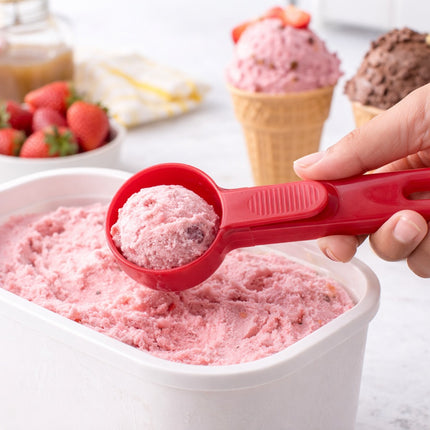Red ice cream scoop taking a portion of pink ice cream from a container, with ice cream cones and strawberries in the background.