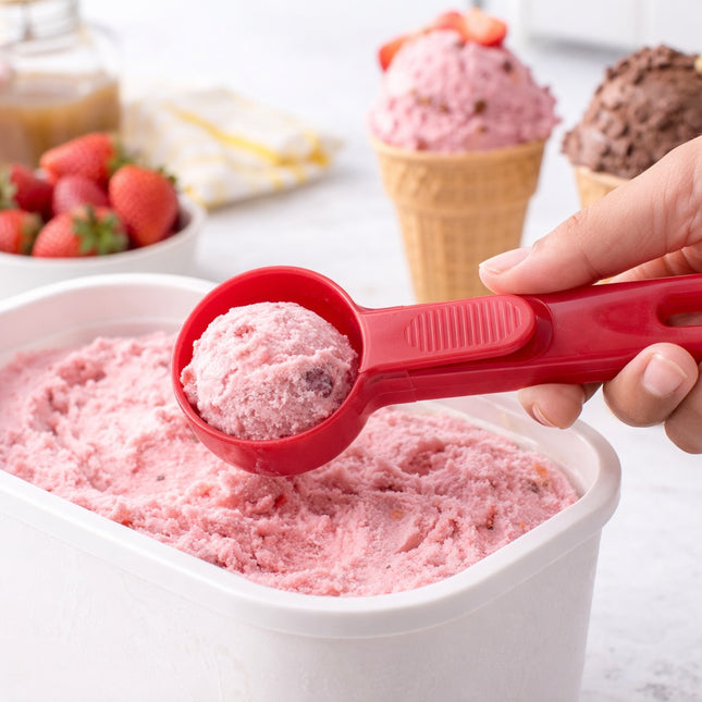 Red ice cream scoop taking a portion of pink ice cream from a container, with ice cream cones and strawberries in the background.