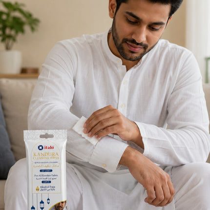 A man in a white kurta sitting indoors applies a wet wipe from a packet of Italo Kandura Classic Wipes to his wrist. The packet is clearly visible in the foreground on a light-colored surface.