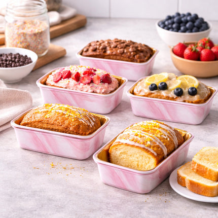Assorted baked goods in pink loaf pans on a light gray surface with fruits and ingredients.
