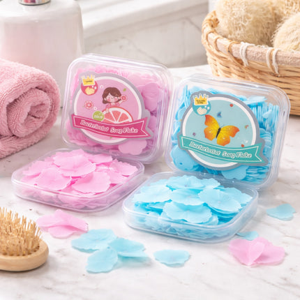 Two containers of pink and blue soap flakes on a bathroom counter with a brush and towel in the background.