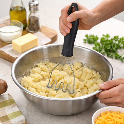 Person mashing potatoes in a bowl with a masher, surrounded by kitchen items.