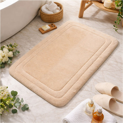 Beige bath mat on a bathroom floor with flowers, soap, and slippers.