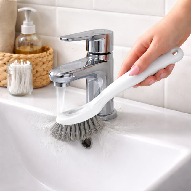 Person using a scrub brush with a handle to clean a sink, with a faucet and bathroom items in the background.