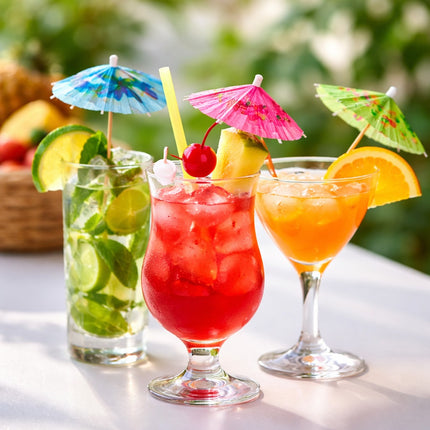 Three colorful cocktails with umbrellas and fruit on a table outdoors.