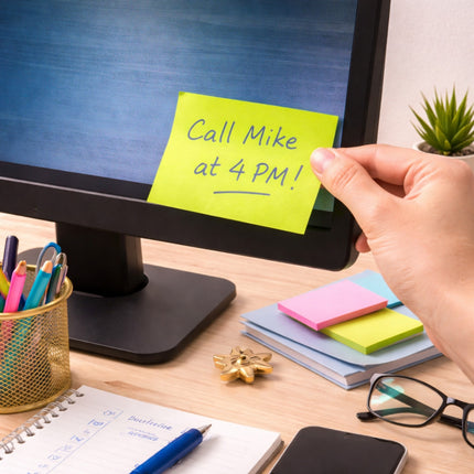 Hand placing a sticky note on a computer monitor with text 'Call Mike at 4 PM!' on a desk.