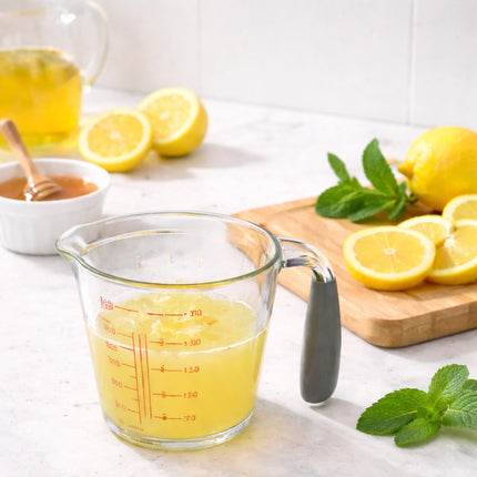 Glass measuring cup with lemon juice, lemons, and honey on a white surface