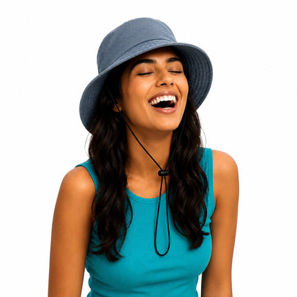 A woman with long dark hair wearing a slate blue bucket hat and a teal sleeveless top, laughing with her eyes closed against a solid white background.