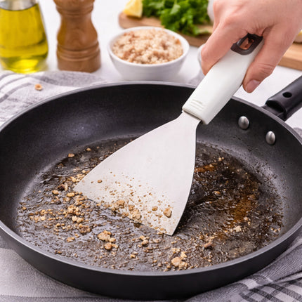 Person using a white spatula to scrape food残渣 from a black frying pan on a kitchen counter.
