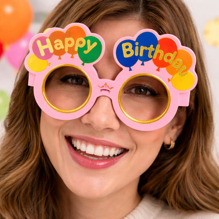 Person wearing colorful 'Happy Birthday' glasses with a blurred festive background