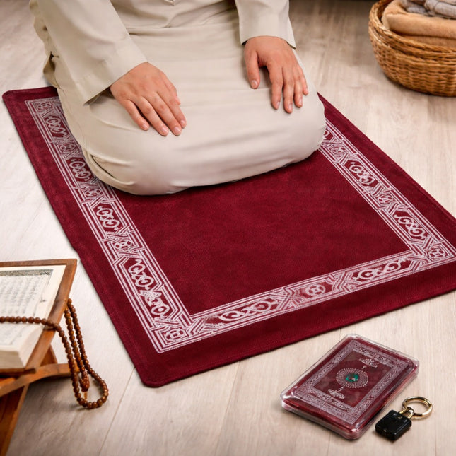 Person praying on a red prayer mat with a Quran and prayer beads on a wooden floor.