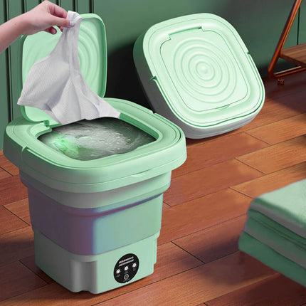 Green collapsible trash bin with a person disposing of a white bag into it on a wooden floor.