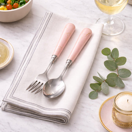 Pink-handled silver cutlery set on a white napkin with a glass of white wine, bowl of salad, and candle in the background.