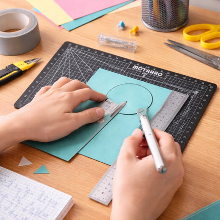 Hands using a craft knife on teal paper with a Motarro cutting mat on a wooden table.