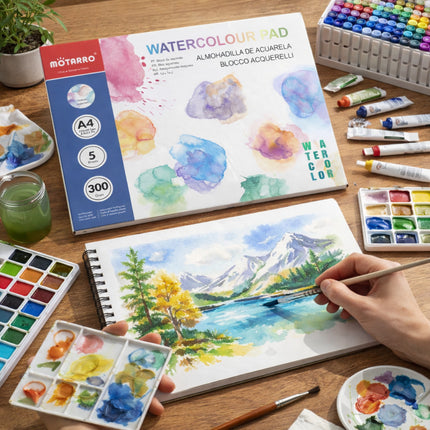 Person painting a mountain landscape with watercolors on a wooden table with art supplies.