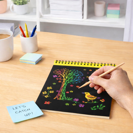 Person coloring a tree and bird design on a black book with colorful markers, on a wooden table.