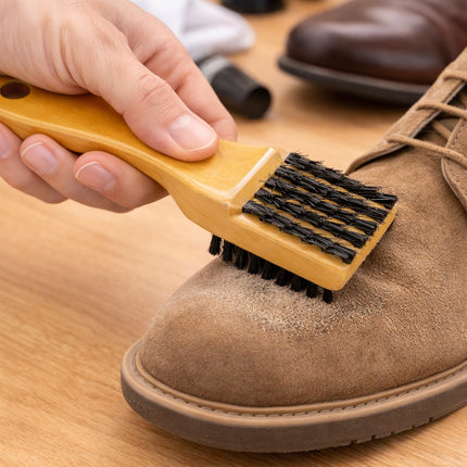 Person cleaning a brown leather shoe with a brush on a wooden surface.