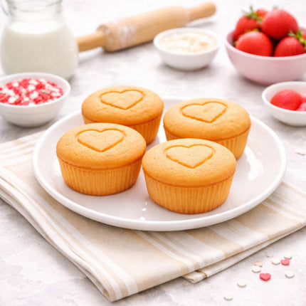 Four cupcakes with heart designs on a white plate, surrounded by baking ingredients on a light surface.