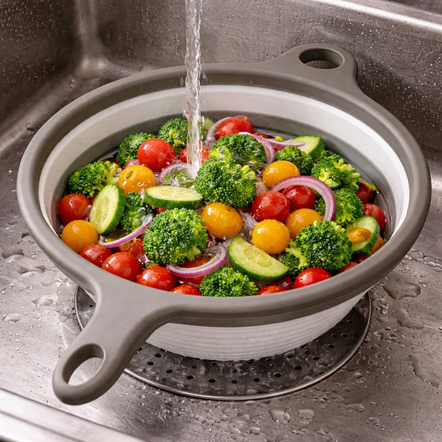 Collapsible colander with vegetables being rinsed under running water in a sink.